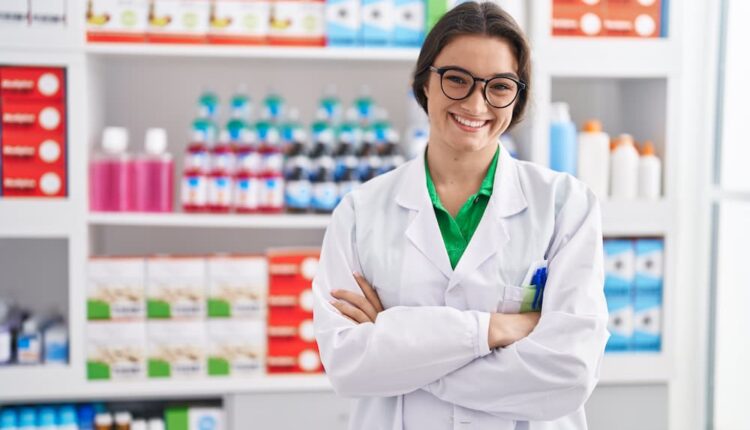 Farmaceutica jovem hispanica sorrindo confiante em pe com gesto de braços cruzados na farmacia
